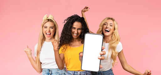 Carefree attractive millennial ladies in stylish casual outfits having fun together on pink studio background, showing smartphone with empty screen, dancing and laughing, mockup, panorama
