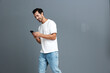 © SHOTPRIME STUDIO - Full body smiling young man wearing white t-shirt and blue jeans, looking at his mobile phone with a cheerful expression against a plain grey background. People lifestyle concept.