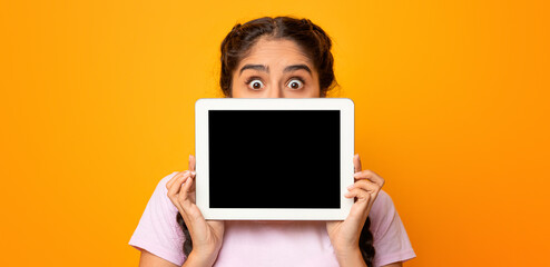 Amazed indian lady hiding behind digital tablet with empty screen isolated over yellow studio background. Excited woman holding and showing pad, mockup, advertisement for application or website