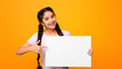 © Prostock-studio - Place For Your Ad. Portrait of smiling indian lady holding empty blank board isolated on orange studio background. Happy woman standing with white square paper for template and pointing at it