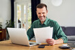 © BusinessClass by W61 - Smiling man working from home using laptop holding documents