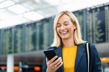 Happy businesswoman using mobile phone in front of departure board
