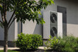 © StockMediaSeller - Modern outdoor heat pump unit installed near a residential house wall, surrounded by green shrubs and a tree in the landscaped yard