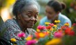 © Riya Kanu - Happy elderly Black African American woman in a wheelchair smelling flowers in a hospice nursing care home garden. African American pensioner with nurse, Generative AI