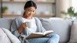 © GrayFrog - Young asian woman relaxes on a comfortable sofa, enjoying a book and a cup of coffee in a cozy home setting.