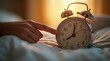© Alex Pios - Hand reaching to stop ringing alarm clock on bedside table in morning bedroom setting