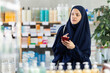 © JackF - Young Muslim woman in traditional islamic navy blue dress and hijab, shopping for medications or care products in drugstore, using phone to check list of needed items