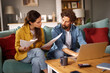 © Stockphotodirectors - Two individuals are seated on a couch, focused on financial documents and a laptop, engaged in a serious conversation about their financial issues.