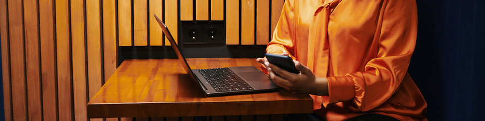 Wall Mural - Header of unrecognizable woman sitting at wooden table using smartphone and laptop, only upper body and hands visible, working or browsing technology in modern workspace