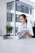 © SOMKID - Smiling Female Doctor with Clipboard and Laptop for Online Medical Consultation working in Modern Clinic.