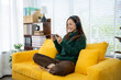 © Kritdanai - Contented Moment: A young woman is absorbed in a mobile game, headphones on, sitting on a vibrant yellow couch within a bright, inviting living space.