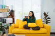 © Kritdanai - Young Woman Gaming on the Sofa: A relaxed young woman engrossed in a mobile game while lounging on a vibrant yellow sofa, immersed in her personal leisure space.
