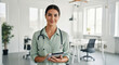 © Chanthakan - Confident female doctor with stethoscope holding digital tablet in modern bright medical office, smiling and looking at camera, professional healthcare worker portrait