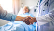 © Rachmadsyah - Doctor shaking hands with a patient's relative in a hospital room, with the patient resting in bed in the background.