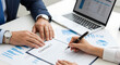 © AWAN - Two business professionals finalizing an agreement by signing a contract document on a desk with financial charts and a laptop.