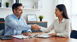 © AWAN - Two smiling business professionals, a man and a woman, shake hands over a desk to seal an agreement.
