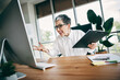 © deagreez - Frustrated businesswoman managing work challenges in a modern office environment, holding a folder and addressing a computer screen