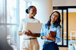 © (JLco) Julia Amaral - Two businesswomen smiling with a laptop and notebook in an office setting