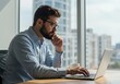 © sujon1638 - Focused professional with beard and glasses intently working on a laptop computer with a city skyline visible through a large window