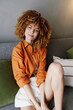 © SHOTPRIME STUDIO - Smiling young woman with curly hair in stylish orange shirt sitting on a couch, showcasing contemporary fashion and joyful lifestyle at home
