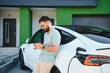 © Serhii - Businessman using smartphone while charging electric car at home