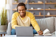 © Prostock-studio - Positive black guy attending online training from home, sitting on couch in front of coffee table with laptop, using headset, taking notes, copy space. E-education, online class, training concept