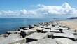 © miss irine - Concrete blocks form breakwater along sandy shoreline under blue sky with white clouds. Calm blue ocean meets coast, with distant lighthouse on horizon. Geometric coastal structure offers protection,