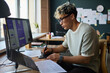 © DragonImages - Young adult Asian man working at desk using digital pen and tablet designing mobile app interface on computer monitor, surrounded by sketches and documents in modern office