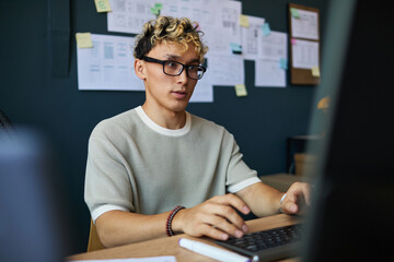 Wall Mural - Young adult man working at computer, sitting at desk in modern office, focusing on screen with hands on keyboard, documents and charts pinned on wall in background