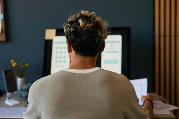 Wall Mural - Young adult Caucasian man sitting at desk working on computer monitor, holding paper in hand, short curly hair with dark and blonde highlights, office workspace visible in background