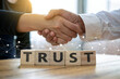 © CatalystVisuals - Closeup of two people shaking hands over wooden blocks spelling out the word trust, symbolizing business partnership and agreement