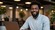 © MstShilpi12 - A smiling african american man with a beard wearing a light blue collared shirt works on a laptop in a modern workspace