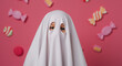 © Wallace - ''A child in a playful white ghost costume with cut-out eyes, set against a vibrant pink background adorned with colorful candi...