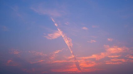 Wall Mural - A timelapse video shows a colorful sunset sky with wispy clouds glowing in soft pink and orange hues against a clear blue backdrop, with a striking diagonal cloud streak adding dynamic contrast.
