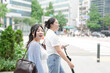 © Masakazu Tokashiki - Two Korean women in their twenties in casual clothes walk along a boulevard sidewalk, talking and smiling as the city awakens under bright spring light, passing shops. Seoul, Jongno District, spring.