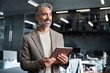 © insta_photos - Smiling middle aged professional businessman manager wearing suit looking away at work in modern space. Happy confident mature business man executive standing in corporate office using digital tablet.