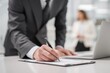 © Padungpong - A business professional in a suit signs a document at a desk with a laptop in a modern office environment