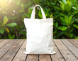 © Deva - White fabric tote bag with handles on a wooden surface, against a blurred green foliage background.