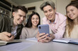 © fizkes - Smart learning with phone. Smiling multiethnic gen z students gather around cellphone at library table watch education content online make group videocall take break from studying to surf social media