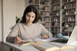 © fizkes - Research before writing. Busy gen z female remote high school student engaged in self training at library desk read paper book use laptop to support written assignment learn information for exam essay