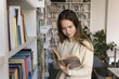 © fizkes - Book lover. Curious young woman pupil student visit public library read literature look through selected volume turn pages with interest search learning information for university degree college essay