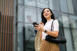 © Parichat - Young businesswoman using smartphone and smiling in front of modern office building