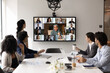 © fizkes - Multinational corporation hybrid meeting. Group of businesspeople top executives from different ethnic backgrounds gather around conference table talk to remote teammates via videocall on big screen