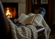 © OneAhmad - A person is reading a book by a fireplace with their feet up, covered by a warm knitted blanket in a cozy and comfortable setting