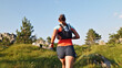 © stockcopter - A woman in athletic gear runs through a scenic, grassy landscape with rocky outcrops and trees. She wears a cap, colorful shirt, and hydration pack, embodying endurance and joy.
