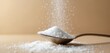 © Viktor - Silver spoon holds white sugar granules against neutral beige background. Minimalist scene fine powder texture, simple culinary element, grainy ingredient. Useful for cooking, baking, pantry concepts.