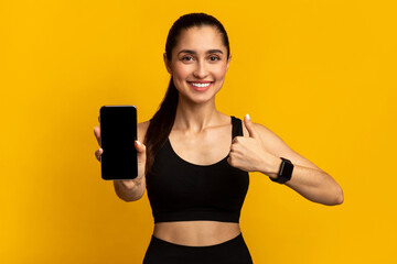 App For Healthy Lifestyle. Portrait of smiling sportswoman holding cellphone with black empty screen for mockup template and showing thumbs up sign gesture, isolated over orange studio background