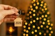 © Chanita - Hand Holding House-Shaped Keychain with Engraving Christmas Tree Background Real Estate Theme Warm Bokeh Lights, Happy New Year 2026 and Christmas Day