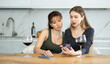 © JackF - Two sad middle-aged women looking at the phone display while sitting at the table with wineglasses at home