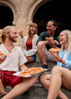 © CarlosBarquero - Vertical. Cheerful young multi-ethnic friends laughing and sharing delicious pizza while sitting on steps in front of a stunning historical building in italy, enjoying a carefree moment together